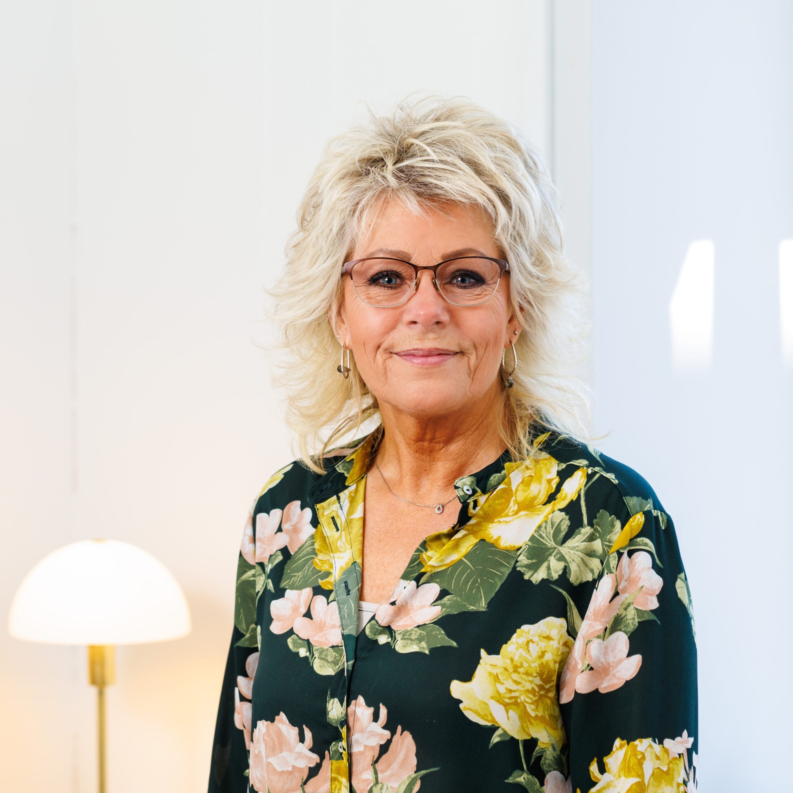 Kvinde med briller og blomsterbluse smiler indendørs